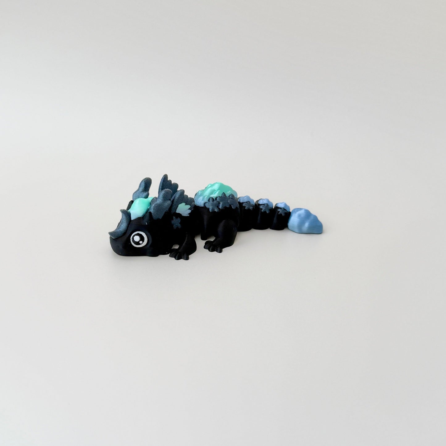 Shimmering black baby dragon fidget toy with starry details, blue-green clouds, and white eyes.